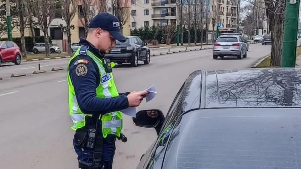 Un polițist în uniformă și vestă reflectorizantă ține documente lângă o mașină neagră pe un drum urban cu clădiri și alte vehicule în fundal.