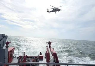 Un elicopter zboară deasupra mării agitate, văzut de pe puntea unei nave care prezintă echipamente de culoare roșie și albă.