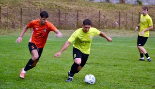 Trei jucători de fotbal sunt pe un teren de iarbă verde, unul cu echipament portocaliu și unul cu echipament galben controlând un balon.