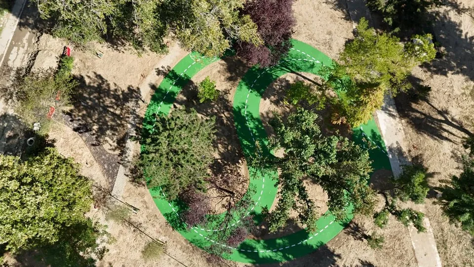 O pistă verde serpentină cu linii albe discontinue trece printre copaci și vegetație într-o zonă cu pământ și iarbă uscată.