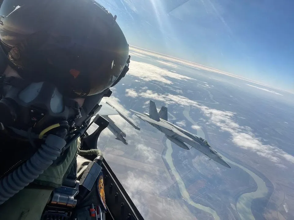 Un pilot cu cască și mască de oxigen privește printr-o trapeză de cockpit către două avioane de luptă care zboară deasupra unui peisaj cu râuri.