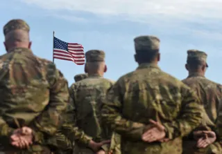 Patru persoane în uniforme militare de tip camuflaj stau cu spatele, privind spre un steag american agitat pe un cer înnorat.