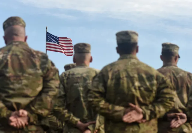 Patru persoane în uniforme militare de tip camuflaj stau cu spatele, privind spre un steag american agitat pe un cer înnorat.