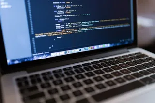 Un laptop deschis afișează linii de cod colorate pe un ecran în întuneric, fiind poziționat în prim-plan pe o suprafață plană.