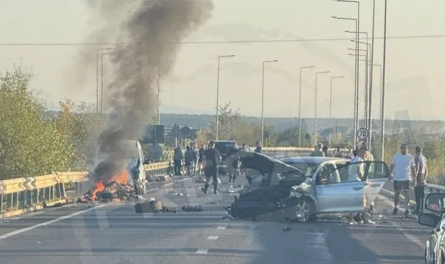Un autoturism argintiu cu capota deschisă stă pe o șosea lângă un vehicul mic care scoate fum negru și flăcări.