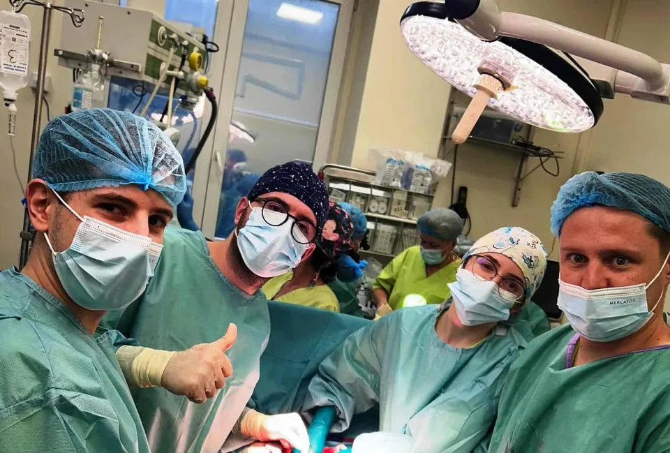 Echipa medicală în sală de operație, purtând mască, cap și halat verde, cu un luminat de operație pe fundal.