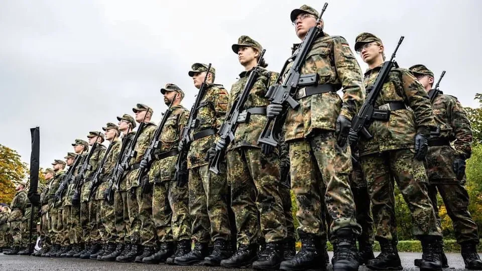 O linie de soldați în uniforme de camuflaj stă în picioare purtând puști de asalt și echipament militar sub un cer înnorat.