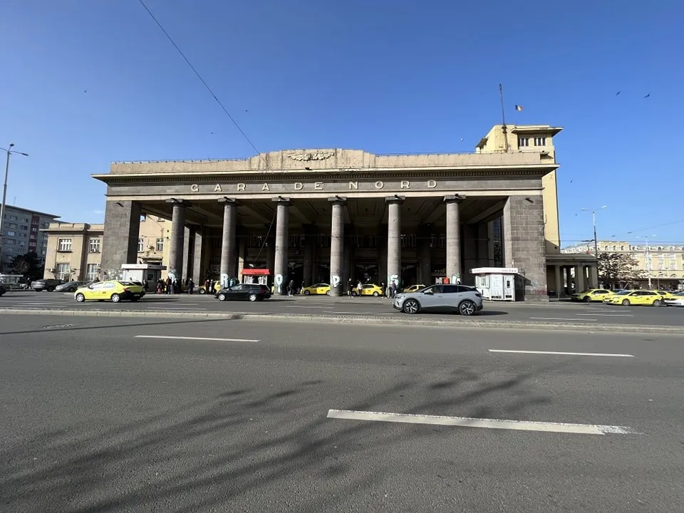 O clădire mare cu coloane susține un acoperiș plat, în fața căreia circulă mașini pe un drum asfaltat sub un cer senin.