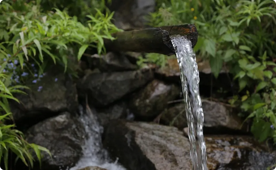 Apa curge dintr-un tub metalic într-un râu de pietre înconjurat de vegetație verde.