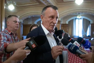 Un bărbat într-un costum închis la culoare este înconjurat de microfoane de la diverse stații de televiziune în timp ce vorbește.