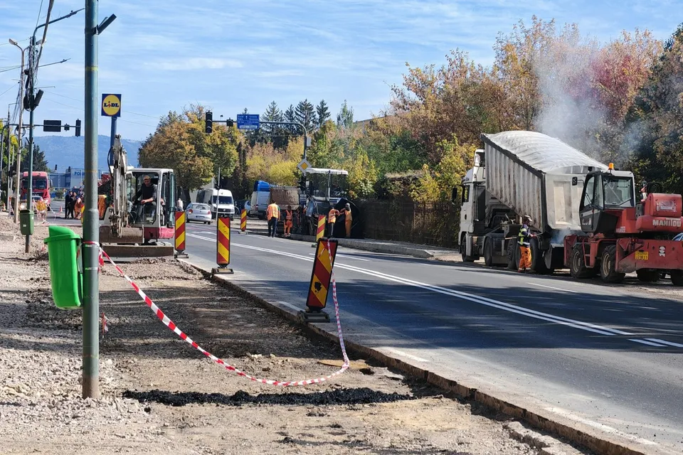 Un camion cu platformă încarcată și utilaje de construcții se află pe un drum asfaltat lângă un trotuar în lucru sub un cer senin.