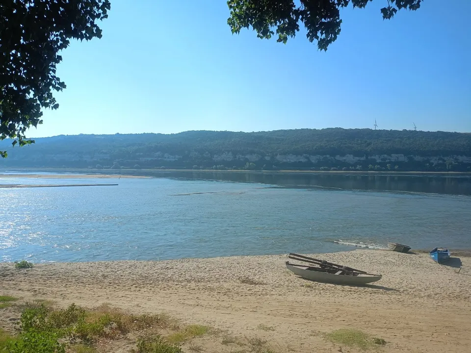 Un râu lat curge sub un cer senin, având o rive nisipoasă în prim-plan cu o barcă mică și vegetație pe margini.