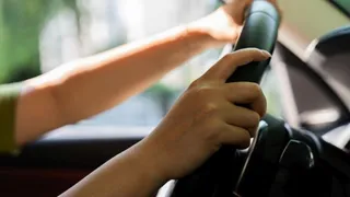 Mâinile unei persoane sunt poziționate pe volanul unui vehicul în timpul condusului.