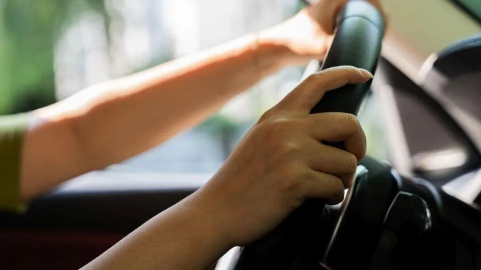 Mâinile unei persoane sunt poziționate pe volanul unui vehicul în timpul condusului.