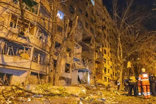 O clădire înaltă cu ferestre sparte și ziduri deteriorate este iluminată noaptea, în timp ce persoane cu veste reflectorizante stau lângă resturi pe sol.