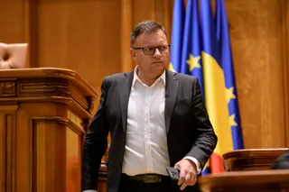 Un bărbat cu ochelari, purtând sacou negru și cămașă albă, stă în fața unor steaguri albastre și galbene într-o încăpere cu mobilier din lemn.