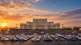 Mulți autoturisme colorate sunt așezate pe o pistă de asfalt în fața unei clădiri mari sub un cer la apus.