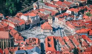O vedere aeriană asupra unui oraș cu numeroase clădiri cu acoperișuri de țiglă roșie și o piață centrală pavată.