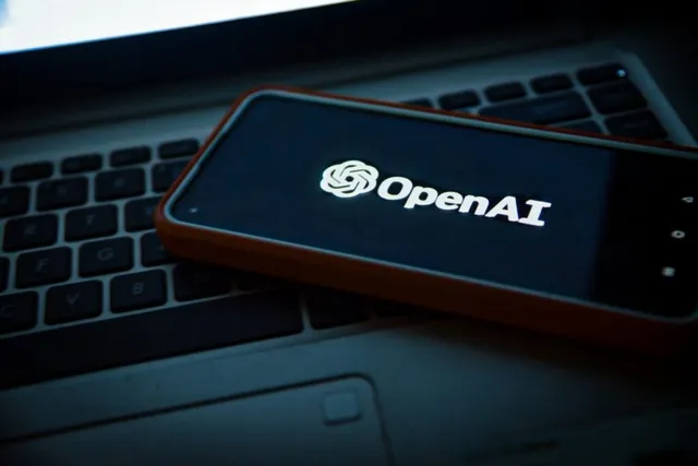 Un telefon mobil cu ecranul aprins afișând logo-ul OpenAI este așezat pe tastatura unui laptop într-un mediu întunecat.