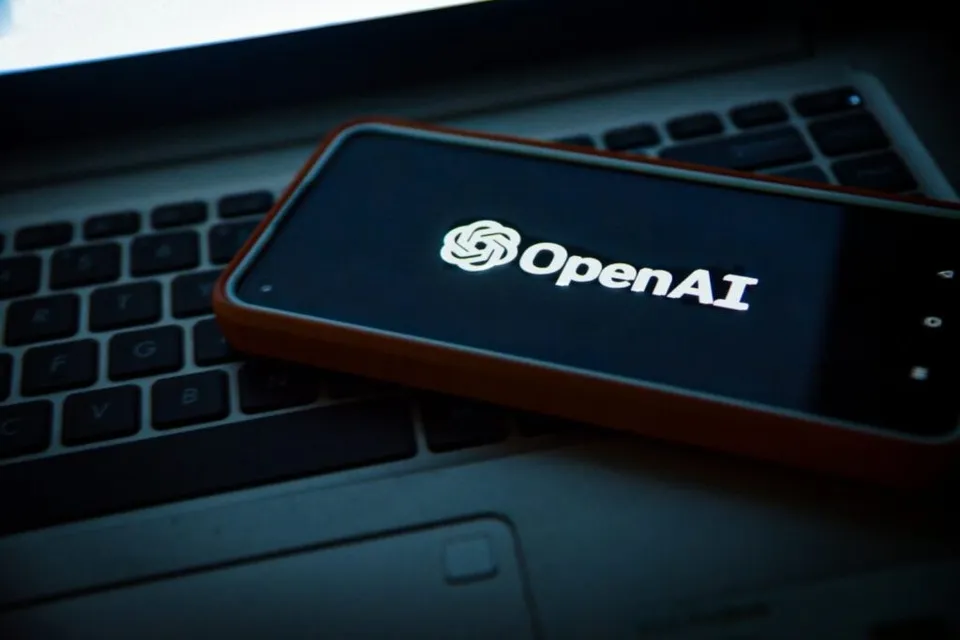 Un telefon mobil cu ecranul aprins afișând logo-ul OpenAI este așezat pe tastatura unui laptop într-un mediu întunecat.