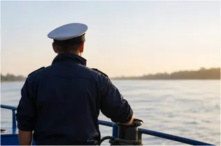 Un bărbat cu șapcă albă și uniformă închisă privește spre un fluviu de pe bordul unei nave.