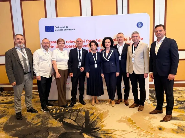 Un grup de nouă persoane stă în picioare în fața unui panou alb într-o încăpere cu podea cu modele maro și galben.