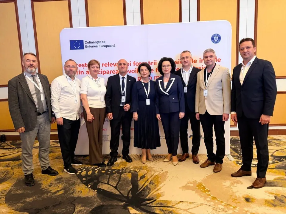 Un grup de nouă persoane stă în picioare în fața unui panou alb într-o încăpere cu podea cu modele maro și galben.