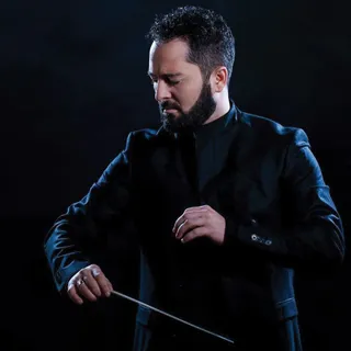 Un bărbat cu barbă și păr scurt, îmbrăcat într-o jachetă neagră, ține o baghetă subțire într-o mână pe un fundal întunecat.