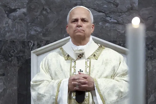 Un bărbat în vârstă cu părul alb, îmbrăcat în haine albe cu broderii aurii, stă cu mâinile împreunate în fața unui perete de piatră gri.