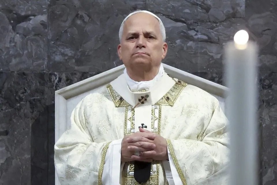 Un bărbat în vârstă cu părul alb, îmbrăcat în haine albe cu broderii aurii, stă cu mâinile împreunate în fața unui perete de piatră gri.