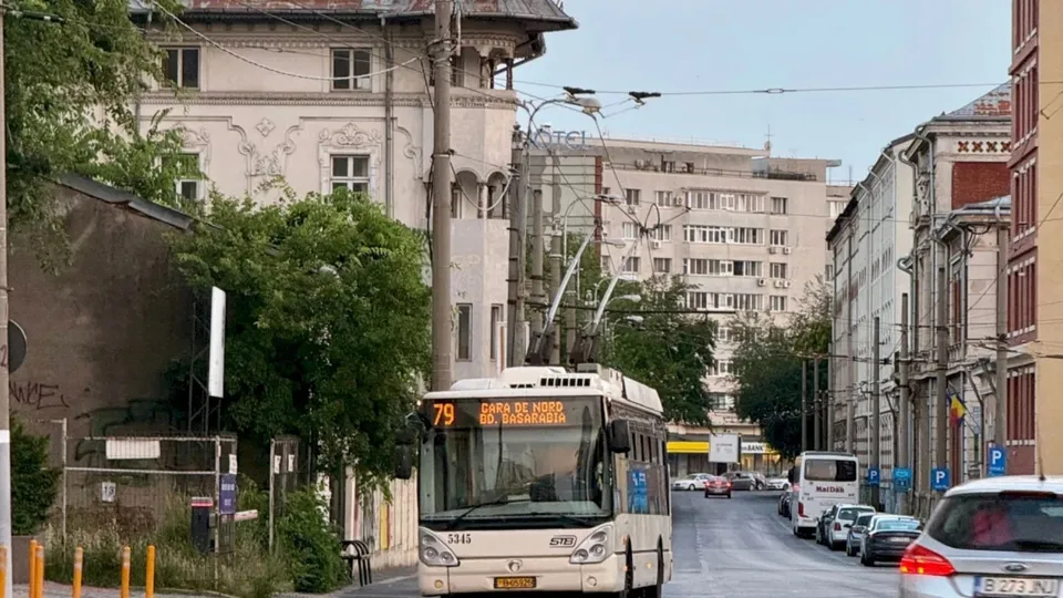 Un trolleybus alb cu numărul 79 circulă pe un drum urban lângă clădiri și copaci sub un cer deschis.