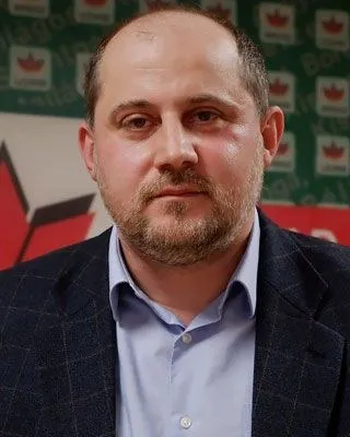 Un bărbat cu barbă scurtă poartă un sacou închis la culoare peste o cămașă de culoare deschisă.