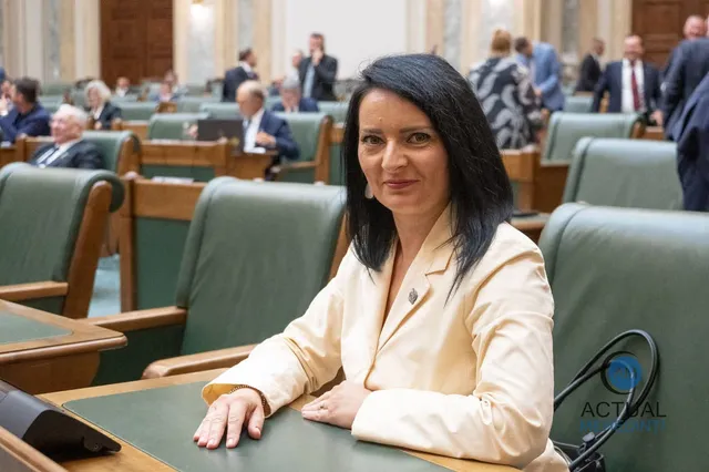 O femeie cu părul negru, purtând un sacou deschis la culoare, stă la o masă într-o sală cu scaune verzi și oameni în fundal.