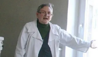 O femeie cu ochelari și halat alb peste un tricou verde stă în picioare lângă o fereastră întredeschisă.