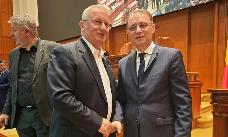 Doi bărbați în costume purtând cravată și ochelari stau unul lângă celălalt într-o încăpere cu panouri de lemn în fundal.