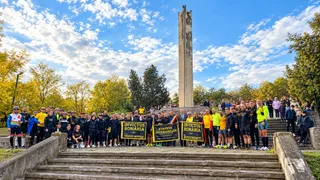 Un grup de oameni stă pe trepte în fața unui monument înalt, purtând haine sportive și ținând trei afișe dreptunghiulare colorate.