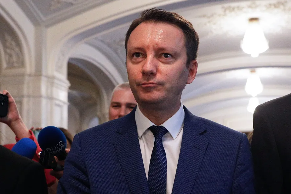 Un bărbat cu părul scurt, purtând sacou albastru, cămașă albă și cravată închisă, stă în fața unor microfoane într-un spațiu cu arhitectură clasică.