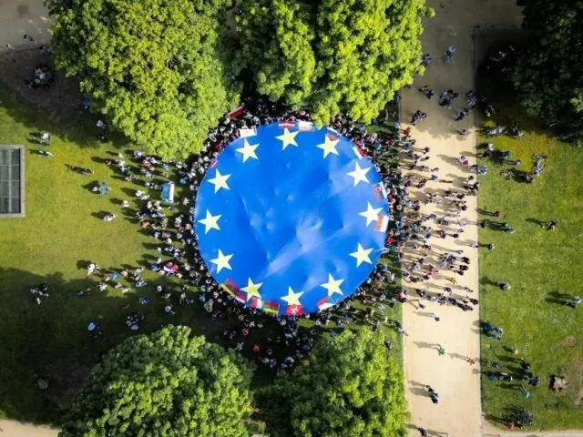 O mulțime de oameni stă în cerc în jurul unui steag albastru mare cu stele galbene pe un teren verde lângă un drum.
