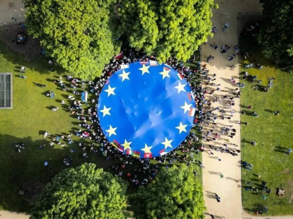 O mulțime de oameni stă în cerc în jurul unui steag albastru mare cu stele galbene pe un teren verde lângă un drum.