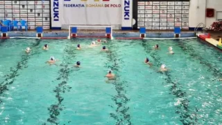 Oameni cu căciuli de înot înoată într-o piscină interioară cu linii negre pe fundul cu apă turcoaz, sub un banner al unei federații de polo.