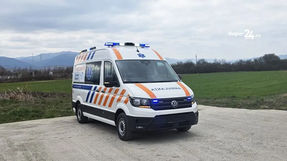 O ambulanță albă cu dungi portocalii este oprită pe un drum lângă un teren înverzit.