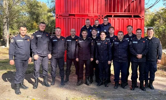 Un grup de oameni îmbrăcați în uniforme de serviciu gri și negre stau în rând în fața unui container roșu.