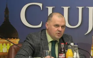 Un bărbat cu părul scurt și sacou gri stă la o masă cu pahare și sticle de băuturi în fața unui fundal albastru cu litere mari.