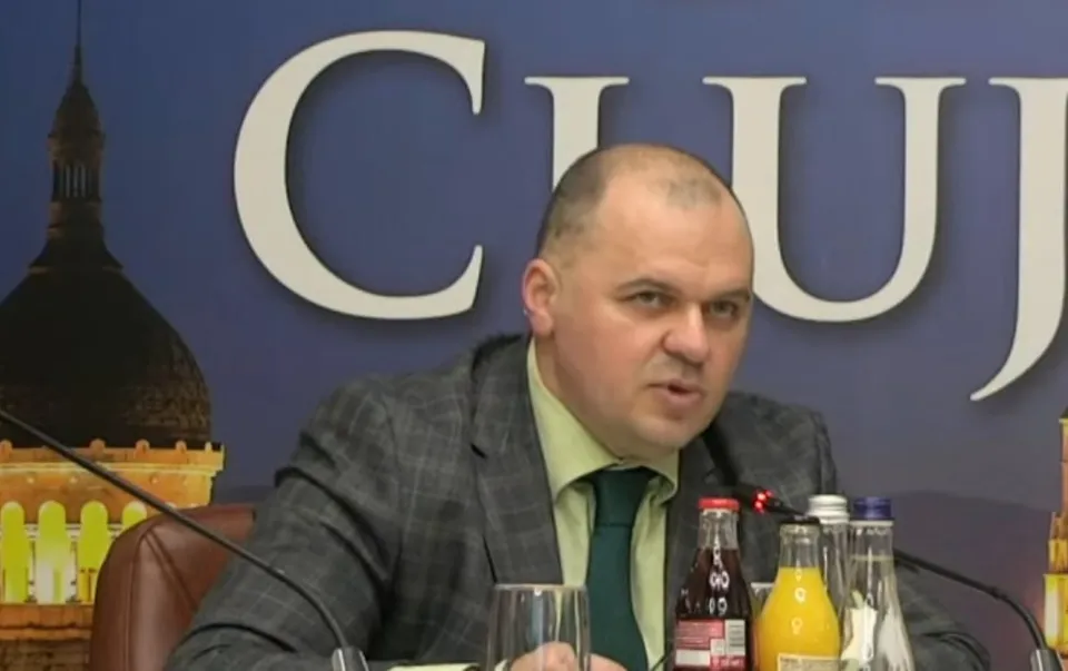 Un bărbat cu părul scurt și sacou gri stă la o masă cu pahare și sticle de băuturi în fața unui fundal albastru cu litere mari.
