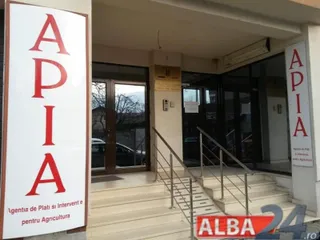 Un panou vertical alb cu litere roșii afișează textul APIA lângă intrarea unei clădiri cu uși de sticlă și scări cu balustrade metalice.