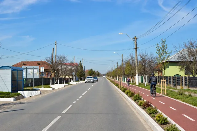 O pistă de biciclete roșie separată de un drum asfaltat este utilizată de două persoane sub un cer senin și luminos.