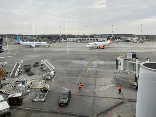 Două avioane sunt parcate pe o pistă de aeroport, lângă echipamente de asistență și două persoane în veste reflectorizante pe asfalt.