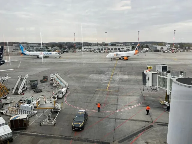 Două avioane sunt parcate pe o pistă de aeroport, lângă echipamente de asistență și două persoane în veste reflectorizante pe asfalt.