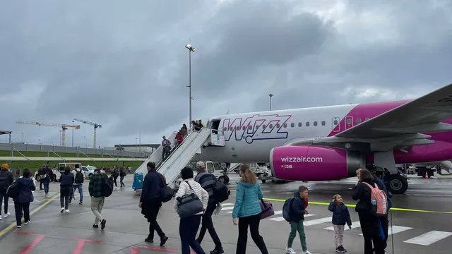 Pasageri cu bagaje merg pe pista unui aeroport lângă o aeronavă roz și albă cu o scară de îmbarcare conectată la ușa din față.