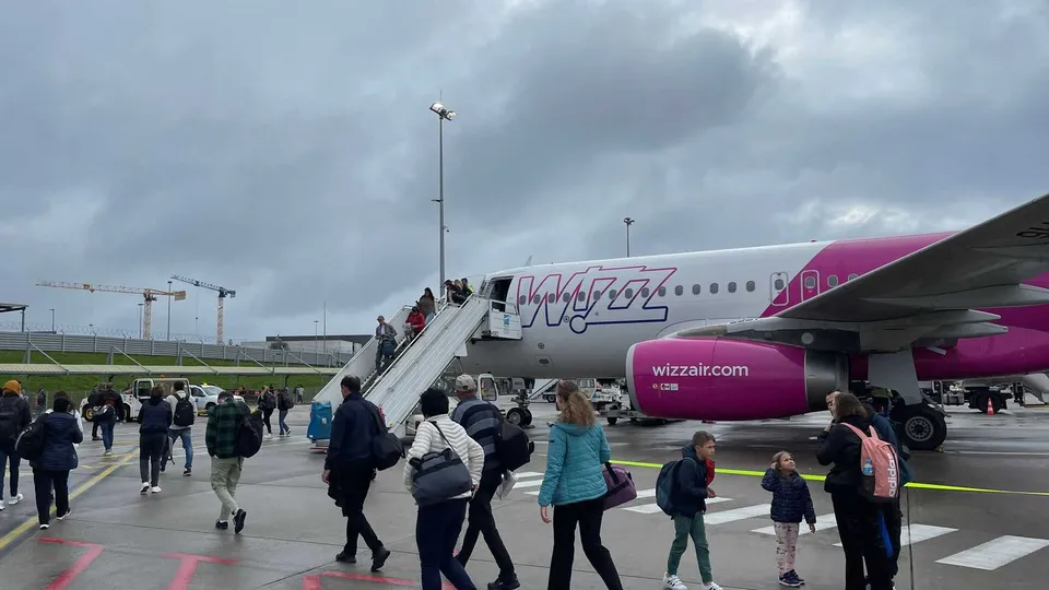 Pasageri cu bagaje merg pe pista unui aeroport lângă o aeronavă roz și albă cu o scară de îmbarcare conectată la ușa din față.